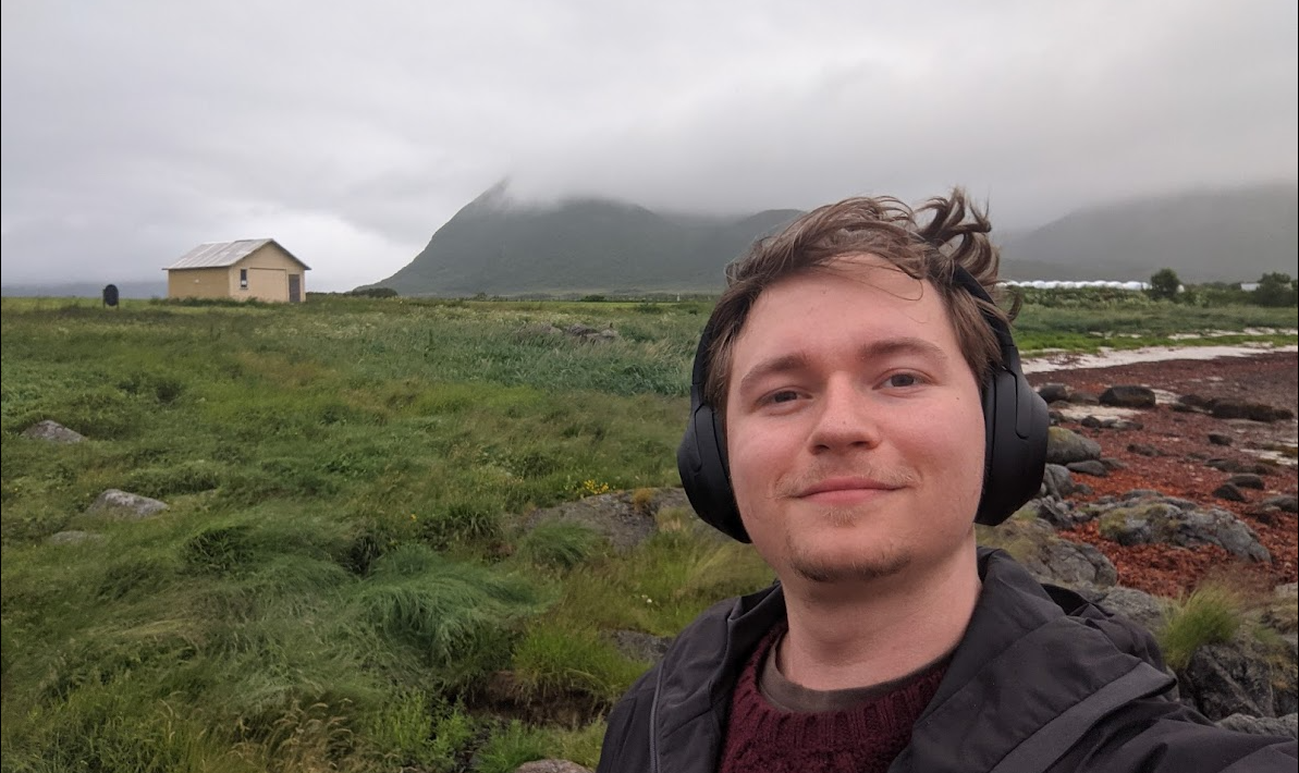 Selfie of me on a windy/foggy day, with a yellow house and a mountain in the background.